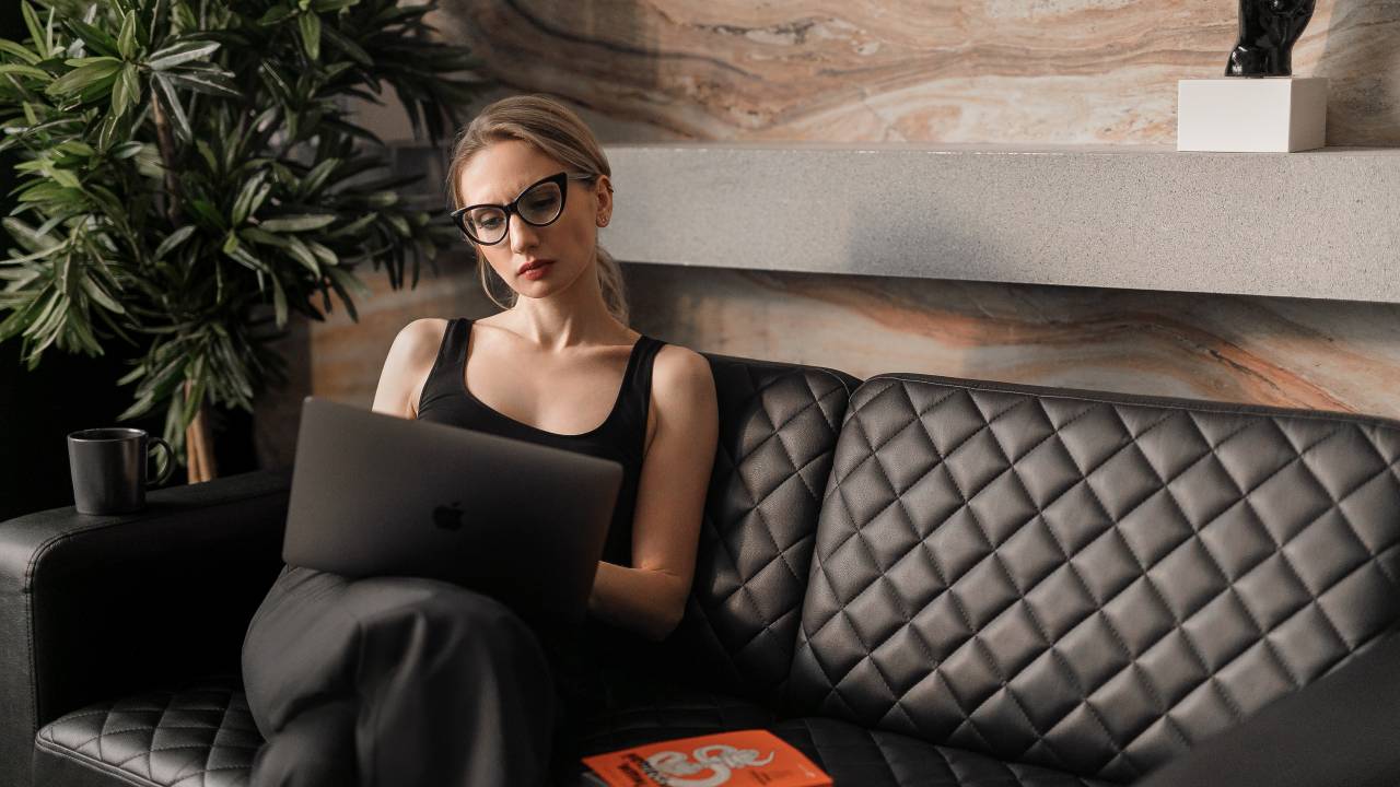 Imagem mostra uma mulher loira, de óculos e cabelos presos, sentada em um sofá com o notebook no colo e um livro ao lado.