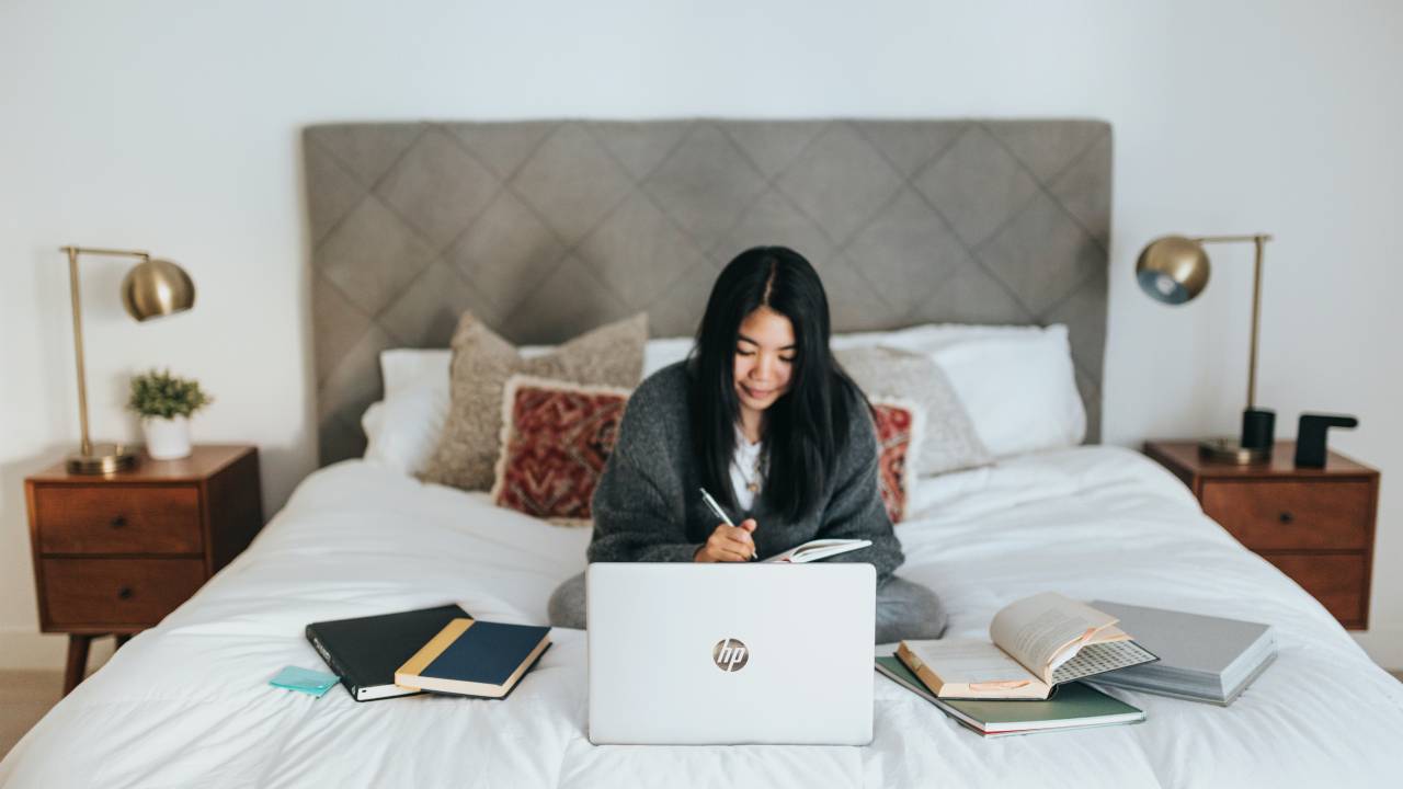 Foto mostra uma mulher oriental sentada na cama em frente ao notebook, anotando algo em um caderno, cercada de cadernos e livros.