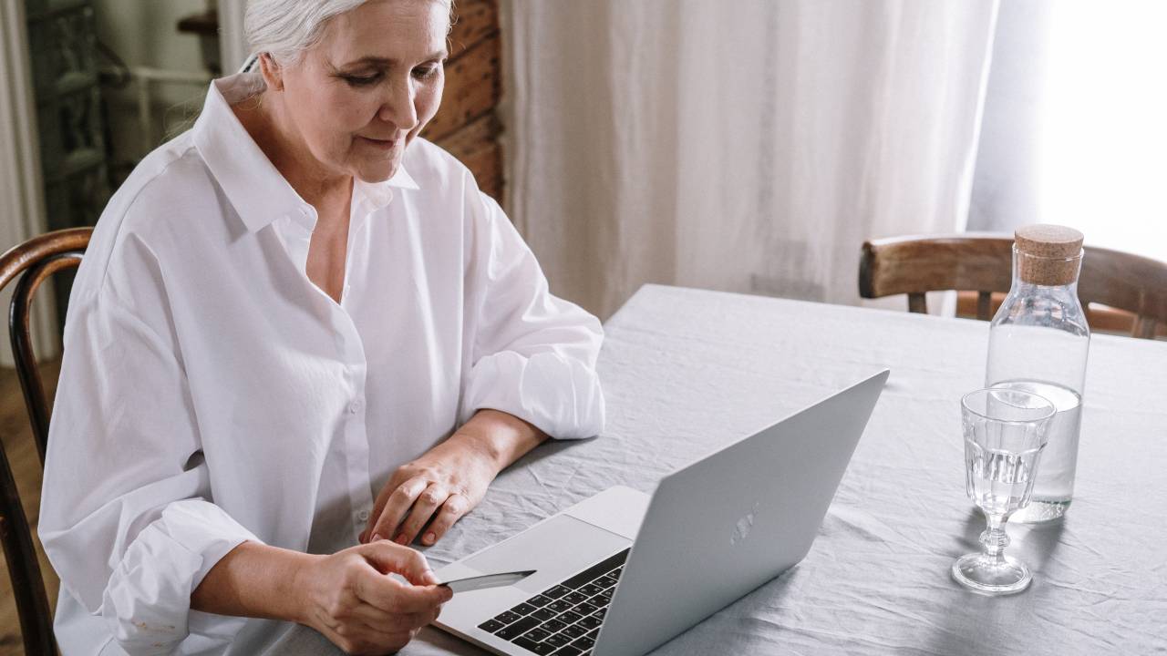 Imagem mostra uma mulher de cabelos grisalhos e camisa branca em frente a um notebook.