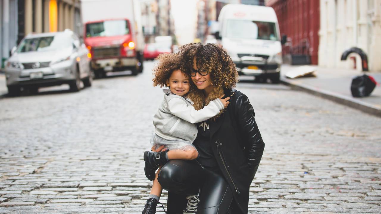 Imagem mostra uma mulher negra de roupa preta abraçando uma menina negra de roupa branca e cinza. As duas estão em uma rua de paraleleípedos com carros ao fundo.