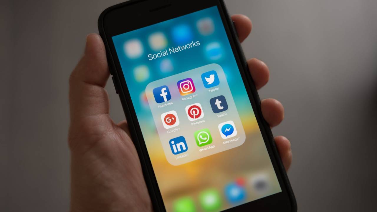 A imagem mostra uma mão segurando um smartphone, na tela estão dispostos ícones de redes sociais