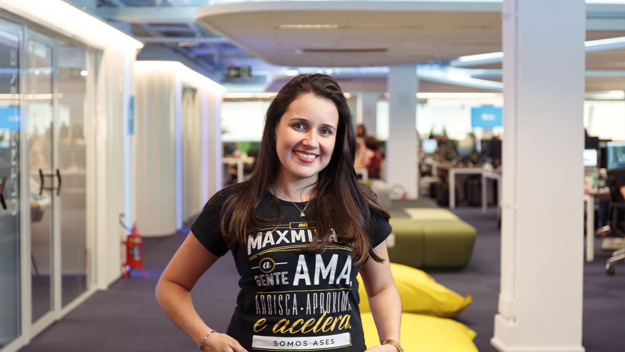 Luiza veste uma camiseta preta da MaxMilhas e está com as mãos no bolso enquanto sorri para foto