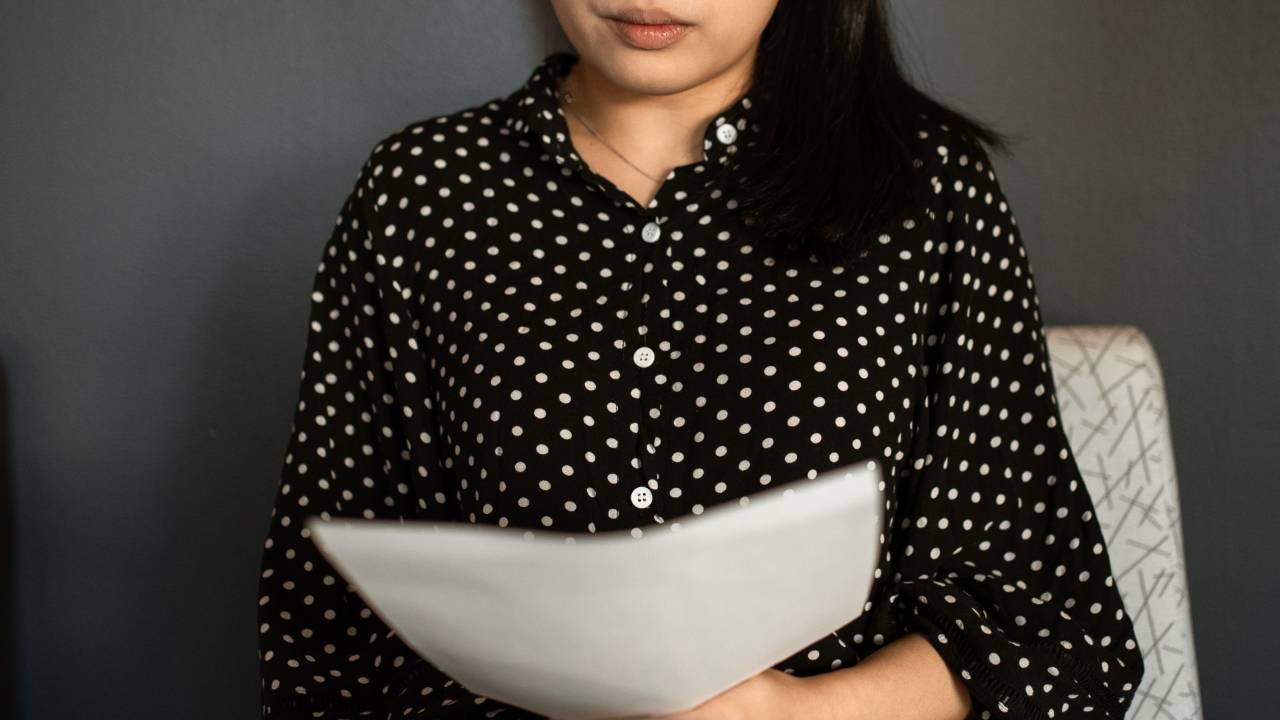 Uma mulher está sentada em uma cadeira, seu semblante mostra apreensão. Nas mãos, ela tem um papel