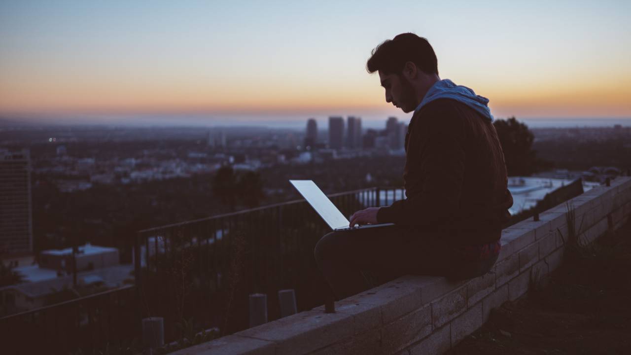 Homem de agasalho com capuz digita em notebook no fim de tarde
