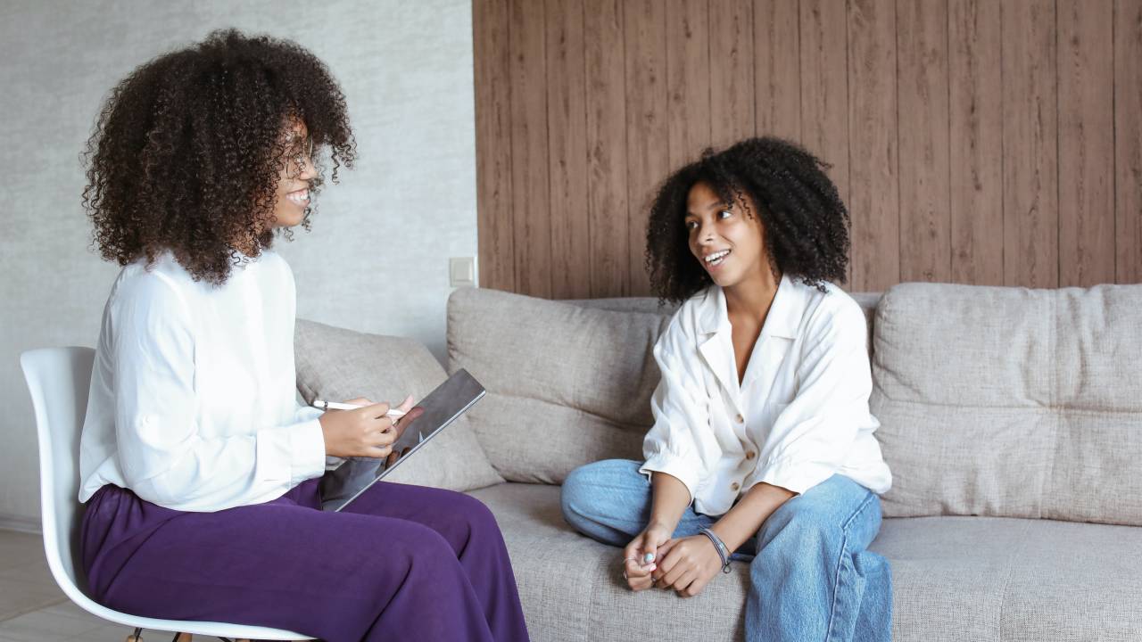 Duas mulheres conversam entre si. Uma delas está sentada de maneira despojada em um sofá, com uma das pernas dobradas. A outra está sentada em uma cadeira.