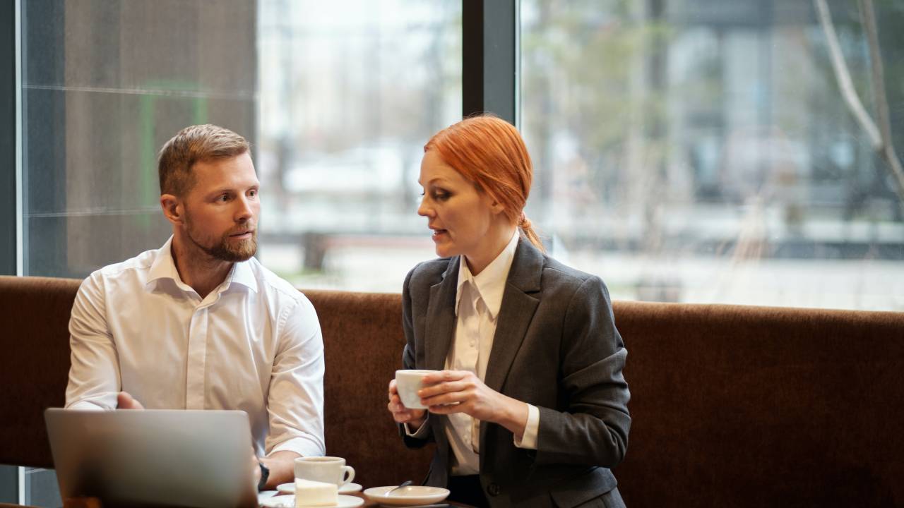 Mulher ruiva com camisa branca e blazer cinza chumbo segura uma xícara de café enquanto conversa com um homem de camisa branca