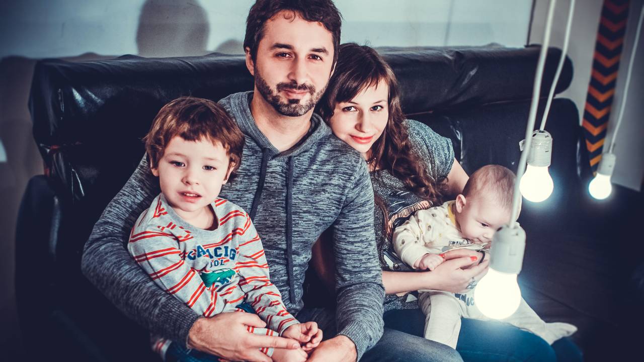 Pai segura criança pequena e mãe segura bebê. Os dois estão no sofá, olhando para a câmera