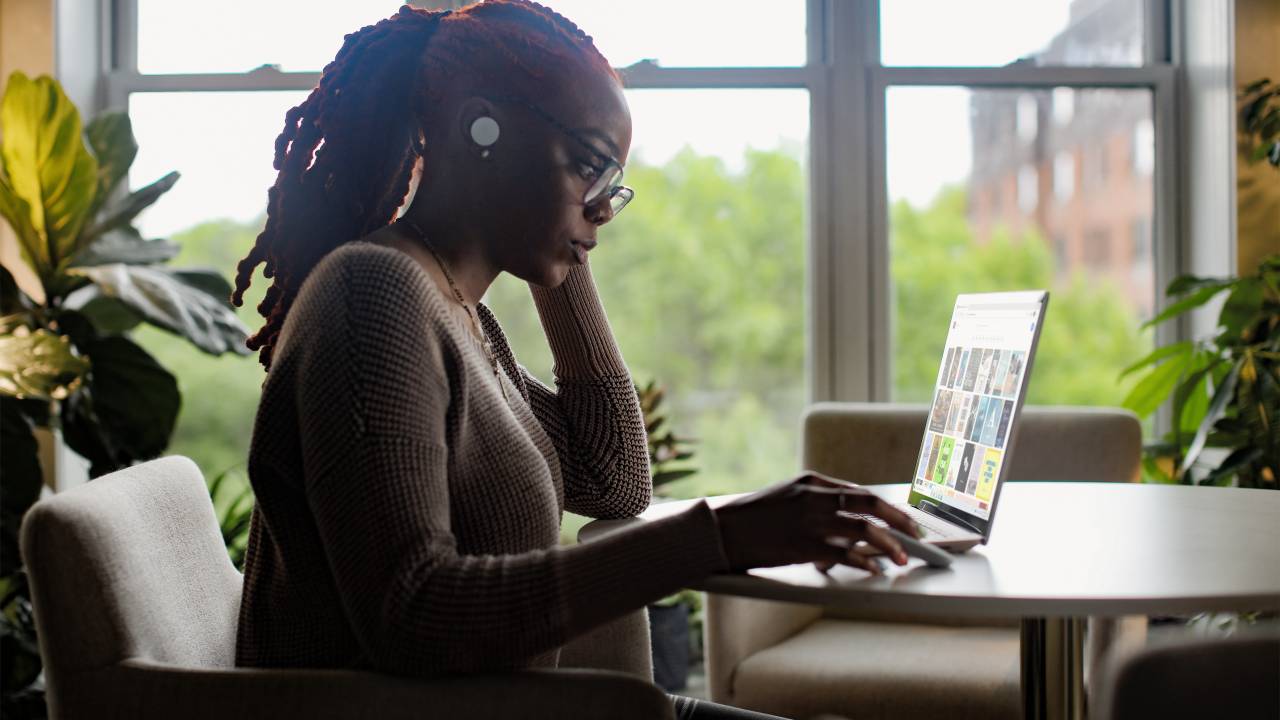 Uma mulher negra está sentada, trabalhando em um computador, ao lado de uma janela. Ela está séria, tem fones sem fio na orelha e usa óculos