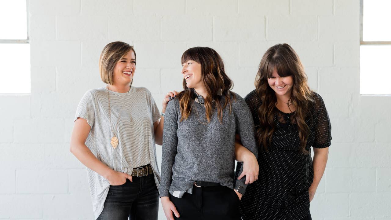 Três mulheres estão de pé, uma ao lado da outra, sorrindo. Elas estão em frente a um fundo branco e duas delas estão com os braços entrelaçados