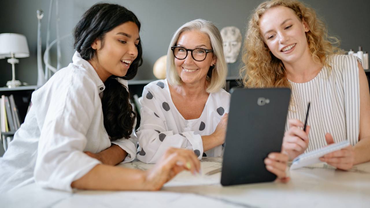 Três mulheres sorriem enquanto trabalham em uma mesa de escritório