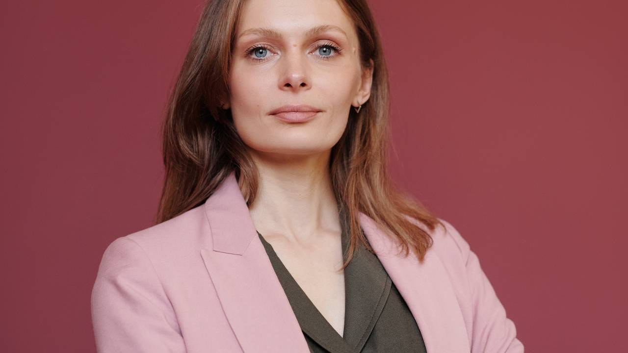 Uma mulher loira veste um blazer rosa claro e cruza os braços