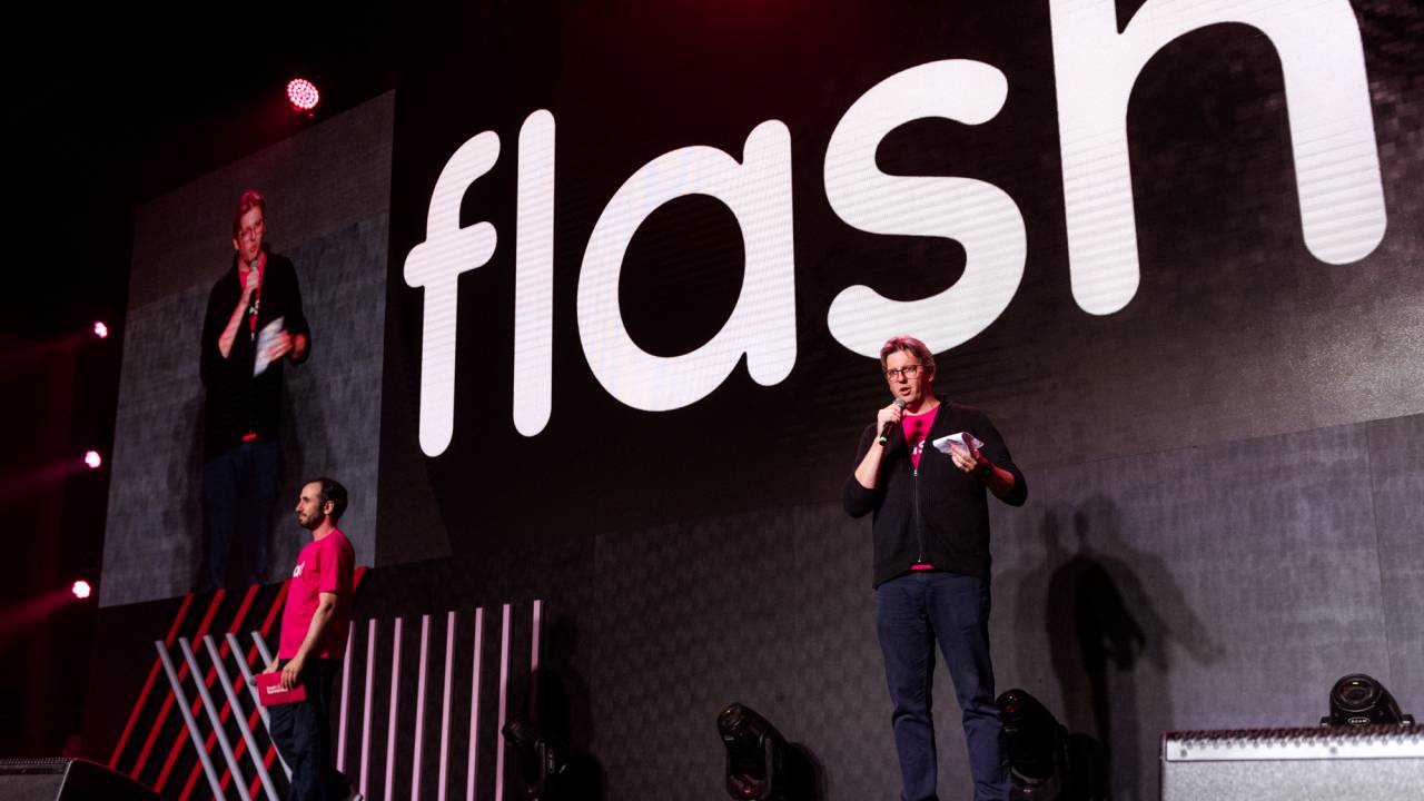 Ricardo e Guilherme estão em um palco com fundo preto e com o logo da Flash em branco