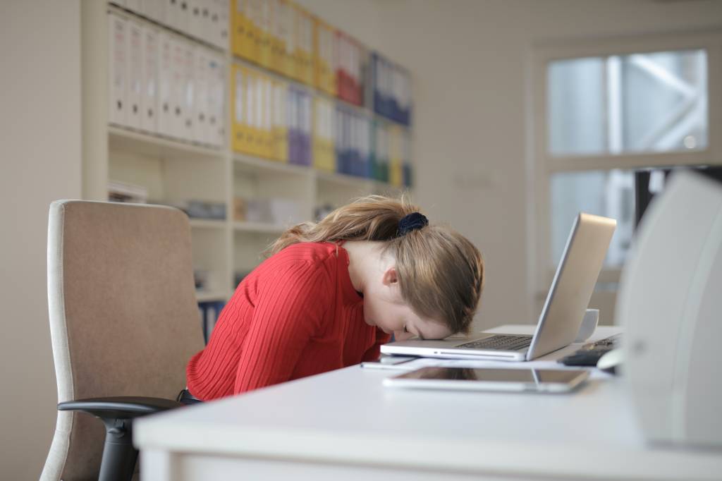 imagem de uma mulher loira em um escritório, sentada à frente de um notebook e com a cabeça apoiada no teclado