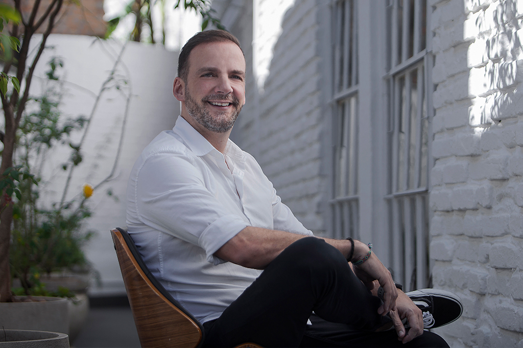 Foto de Raul Aparici, um homem branco com barba grisalha. Ele está sorrindo, sentado em uma cadeira, de pernas cruzadas, e usa camisa branca.