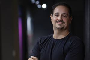 Um homem de cabelos escuros, barba e bigode vestido uma camiseta preta. Ele está de braços cruzados.