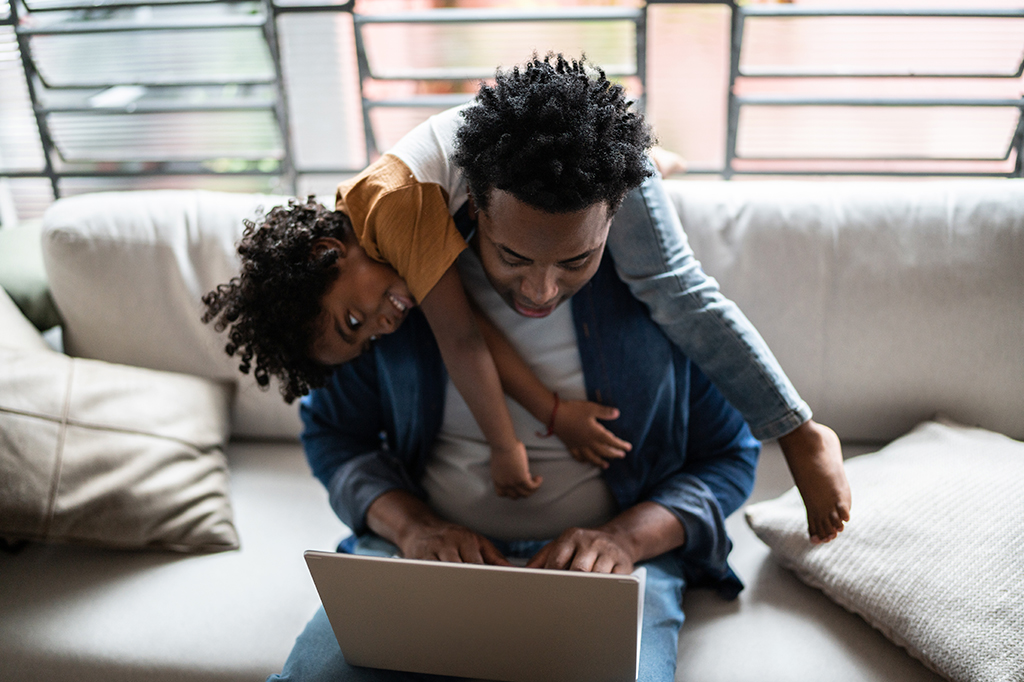 Foto de um pai usando o laptop tentando trabalhar enquanto o filho está nas suas costas brincando.