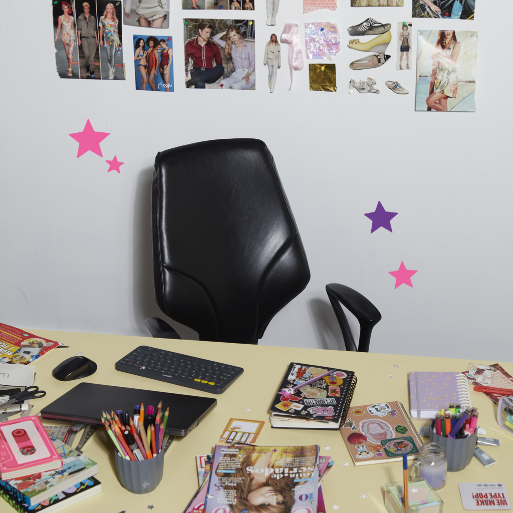Imagem de uma mesa de trabalho, com um notebook, recortes de papel, dois porta lápis e revistas. Ao fundo, uma cadeira vazia e diversas fotos coladas na parede.