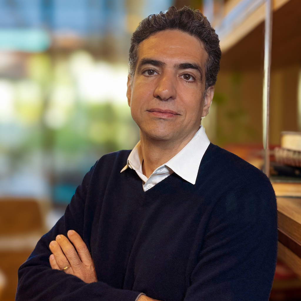 Imagem de um homem, de cabelos pretos, com os braços cruzados, vestindo uma camisa branca e malha azul marinho.